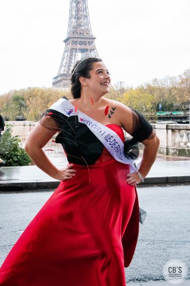 Femme en robe rouge posant près de la tour Eiffel, avec une écharpe "miss curvy île-de-france 2024"