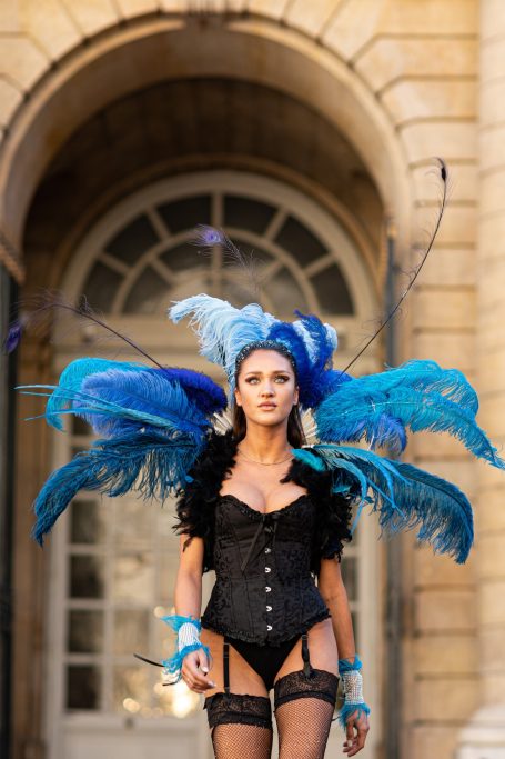 Femme en corset noir avec structure bleu vif et plumes colorées, marchant avec confiance.